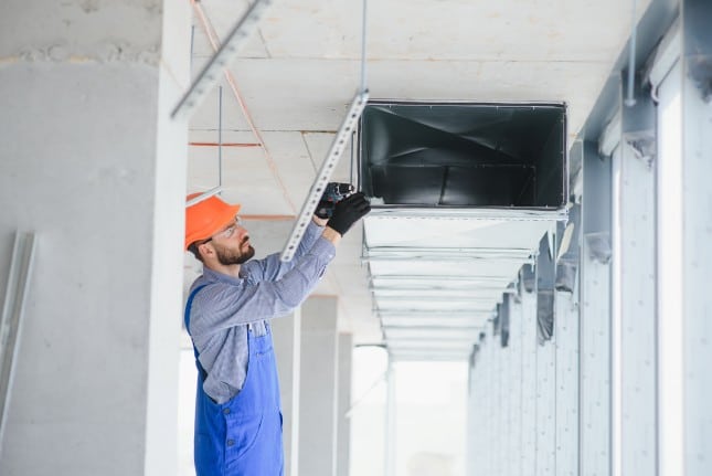 Worker installing or adjusting an air duct in a building under construction, using power tools and wearing safety gear.