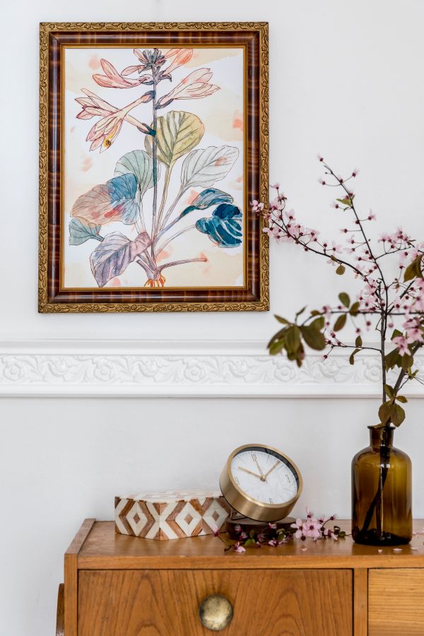 An artistic framed painting of a plant next to a vase with pink flowers, a clock, and a decorative wooden box on a wooden cabinet.