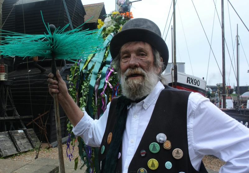 An elderly man dressed in traditional chimney sweeper attire, holding a chimney sweeping tool with eco-friendly bristles, standing in front of a decorated backdrop.