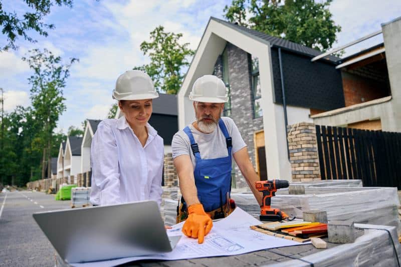 Two professionals, a man and a woman, are reviewing construction plans on a laptop and blueprints at a building site, wearing hard hats and gloves.