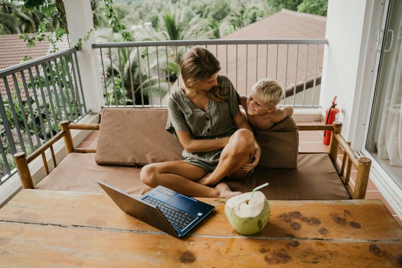 A woman and child relaxing in an outdoor seating area, with a laptop and coconut drink, reflecting a cool and comfortable summer setting.