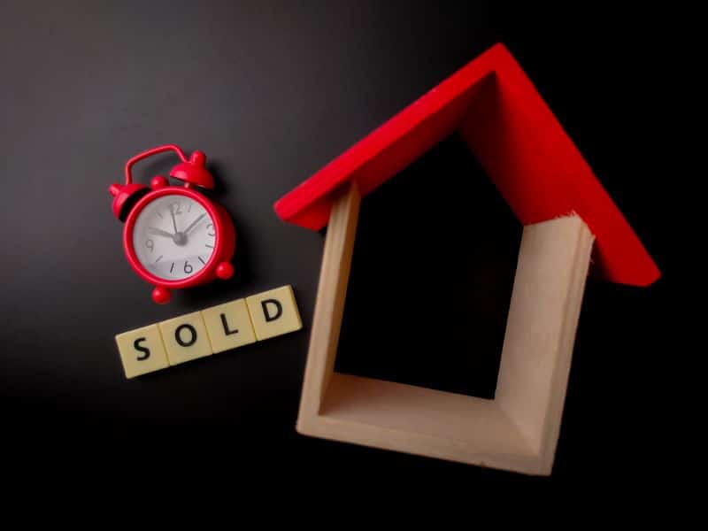 A red alarm clock beside a wooden house model with the word "SOLD" spelled out using letter tiles, symbolizing a quick home sale.