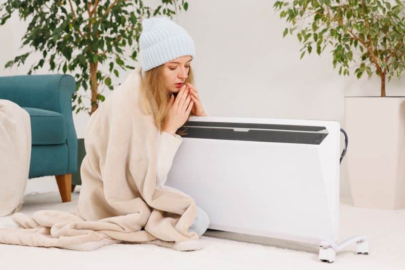 A woman wrapped in a blanket sitting near a heater, feeling cold in a cozy home environment.