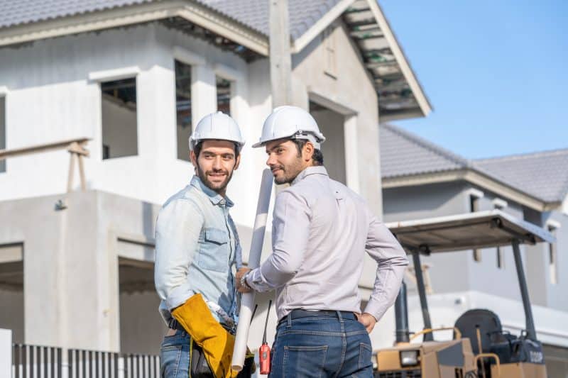 Two construction workers in hard hats and work clothes are posing in front of an under-construction house, holding blueprints and smiling.