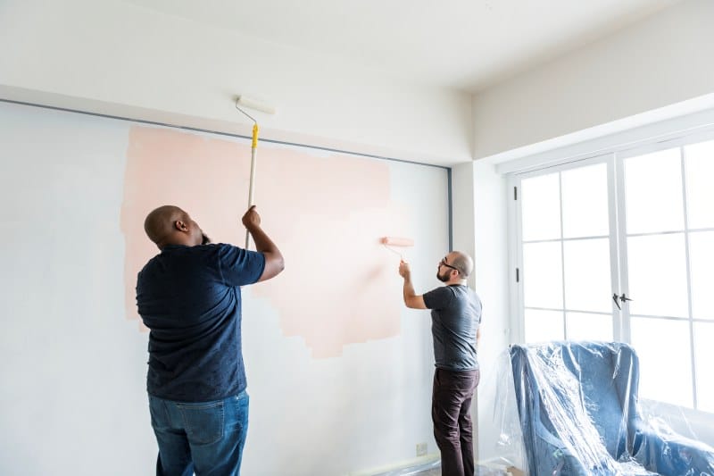 Two people painting a wall in a living room, one using a roller on the top part of the wall, and the other painting the lower section.