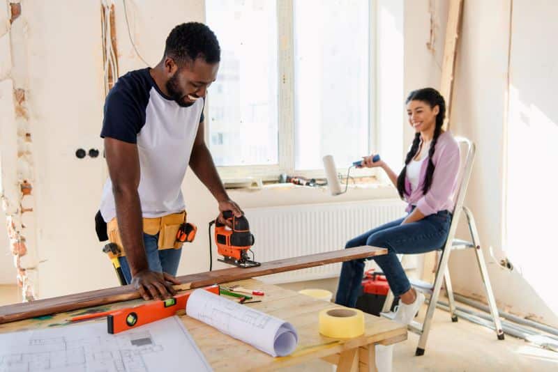 A man working on a wood project with a jigsaw, while a woman smiles and prepares tools for the home improvement project.