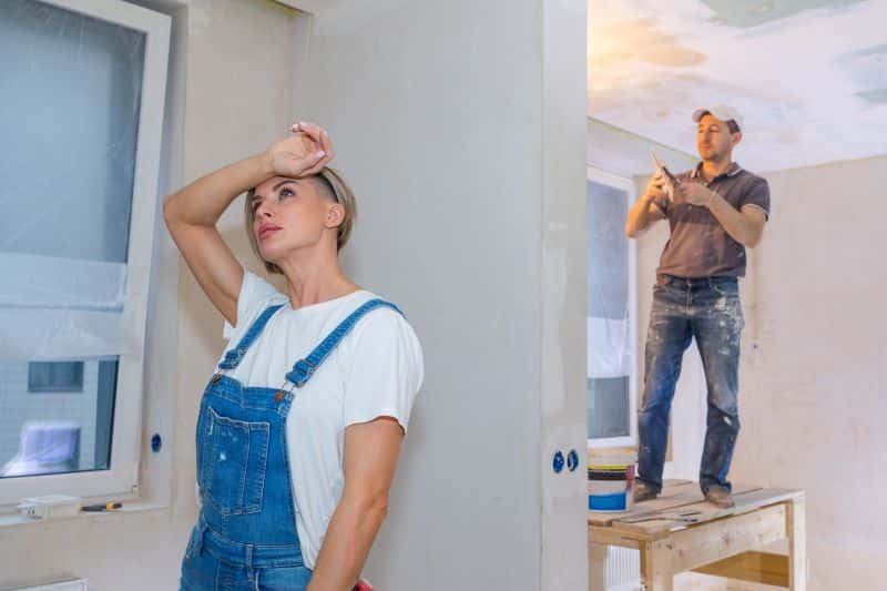 A woman looks stressed while a man works on home restoration, highlighting the challenges of DIY projects and the importance of professional help.