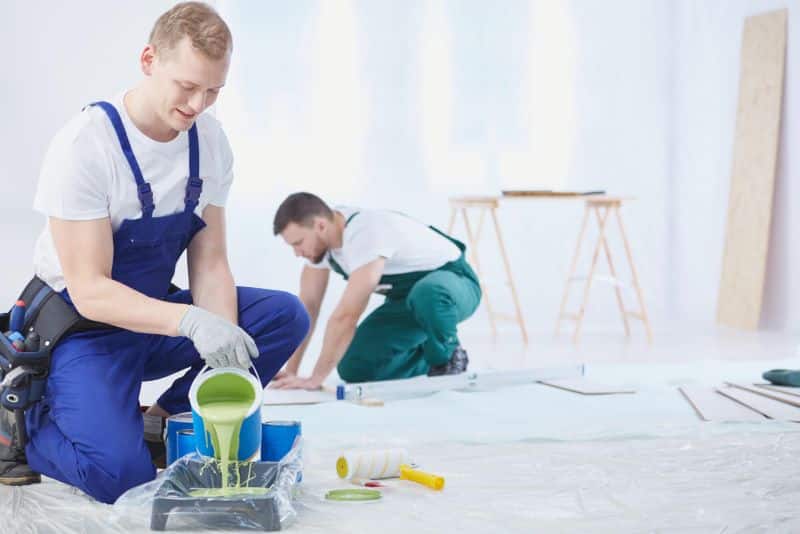 Two workers painting a room, with one pouring paint into a tray and the other preparing the space.