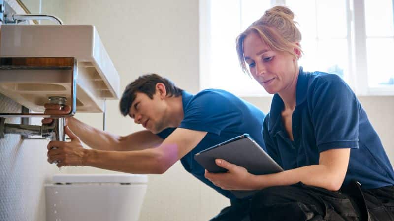 Plumbers performing a plumbing inspection, one working under the sink and the other reviewing details on a tablet, ensuring thorough checks for potential issues in the plumbing system.