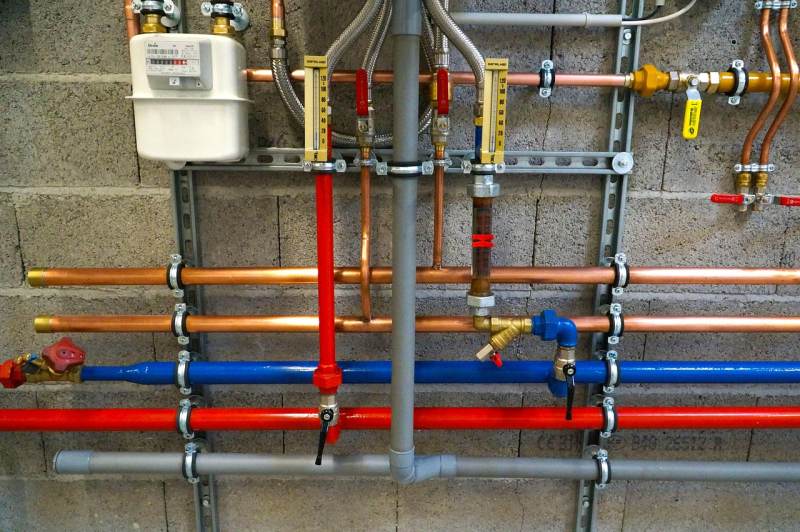 A close-up image showing various pipes and plumbing connections, including copper, blue, and gray pipes, installed on a wall.