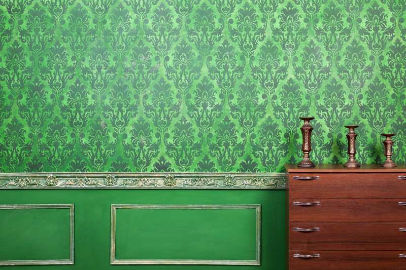Green floral patterned wallpaper with ornate molding at the base and a wooden dresser with decorative candlesticks placed on top.