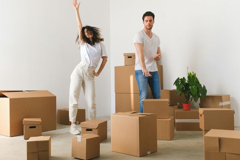 Young couple surrounded by cardboard boxes, smiling and dancing during a local house move, suggesting a budget-friendly and stress-free relocation.