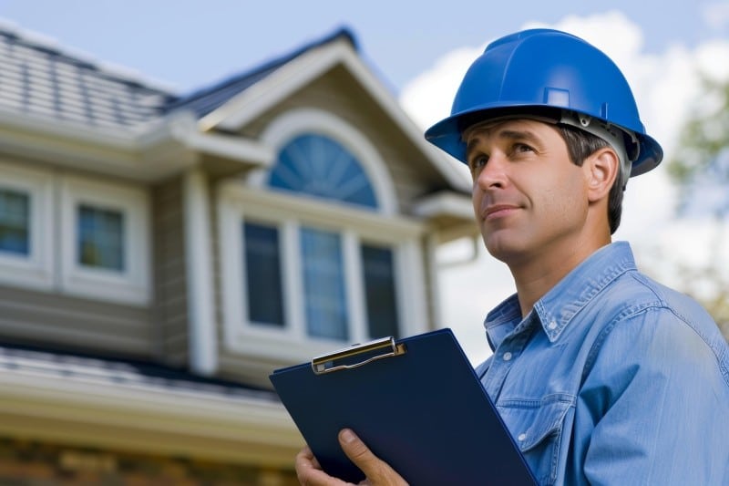 Licensed general contractor in blue hard hat inspecting a residential property with clipboard, assessing permitting requirements for construction compliance.