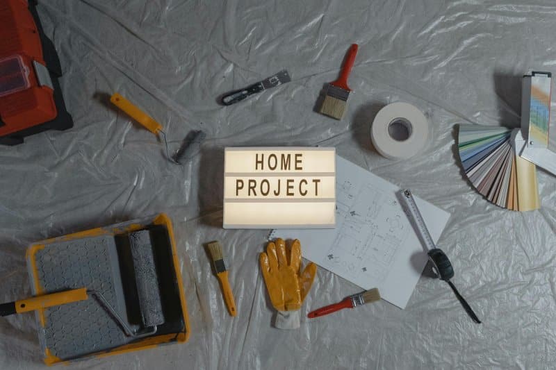 Overhead view of a DIY home renovation setup with tools including paint rollers, brushes, gloves, tape, a toolbox, and blueprints on a plastic-covered floor, featuring a lightbox sign reading “HOME PROJECT”; includes visible safety gear items like protective gloves.