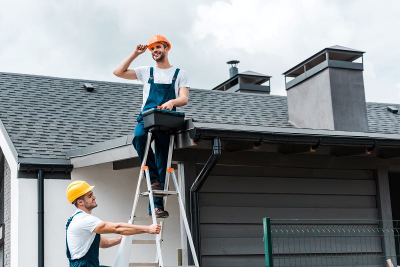 Two roofers in safety gear replacing a shingle roof on a house in Hendersonville, NC using a ladder and tool box.