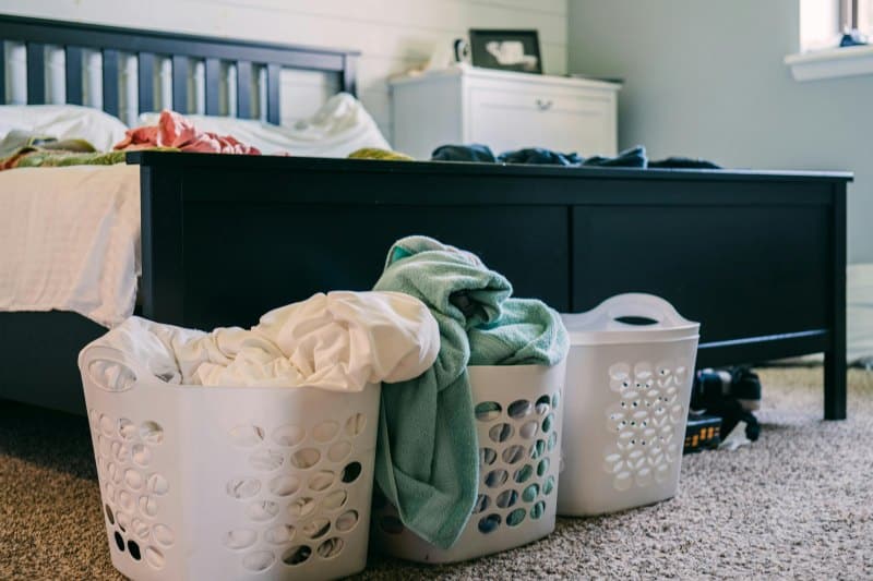 Three laundry baskets filled with clothes placed at the foot of an unmade bed in a cluttered bedroom, illustrating the need for transforming spaces through home decluttering.