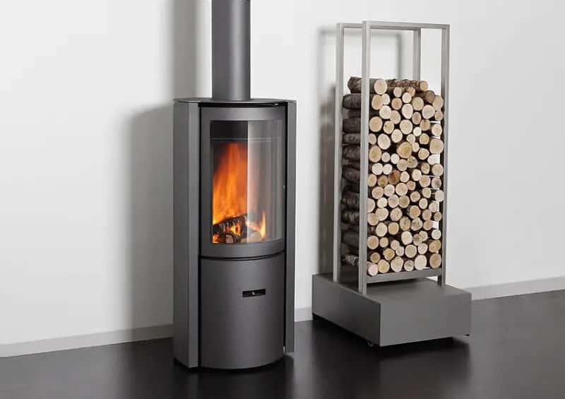 A modern wood stove with a large glass window displaying a natural fire, paired with a metal log holder filled with firewood.