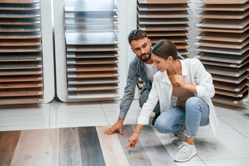 A man and a woman are inspecting flooring samples in a store. The woman is kneeling down, pointing at a wooden floor sample, while the man stands behind her, looking at it. There are various wooden planks displayed on shelves in the background.