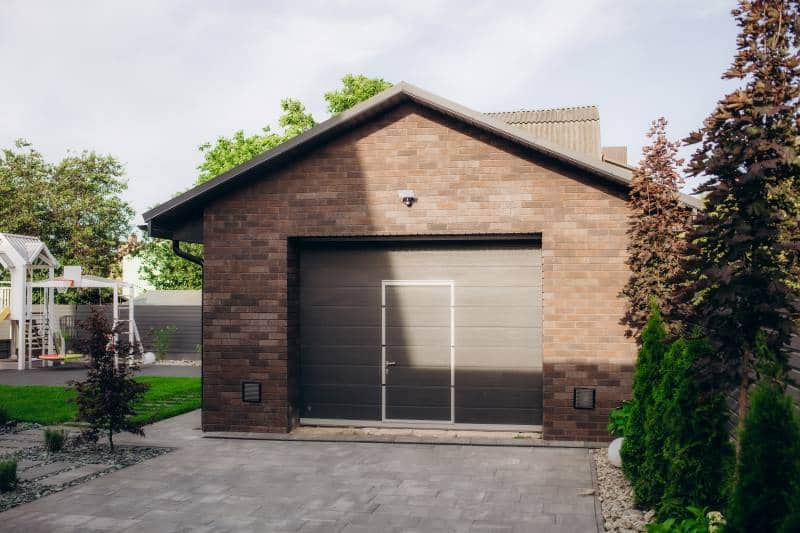 A modern brick house with a metal garage door, surrounded by greenery, including shrubs and trees.