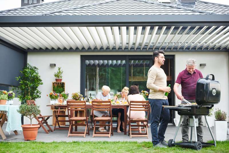 A group of people enjoying a barbecue in the backyard, with one person grilling and others sitting around a table. Plants and greenery are placed around the area, and the setting has a modern, cozy vibe perfect for outdoor living.