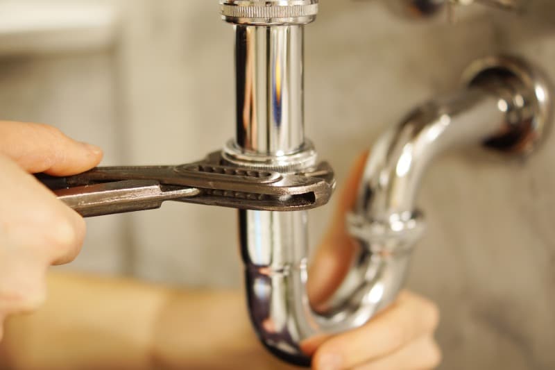Plumber inspecting plumbing system under kitchen sink with tools and replacement pipe in hand.