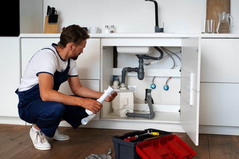 Plumber inspecting and holding pipe under kitchen sink while checking the plumbing system, with a toolbox on the floor.