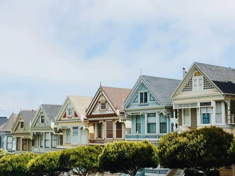 A row of colorful, well-maintained Victorian-style homes with distinct architectural features, situated along a street with manicured shrubs and trees. The image illustrates diverse real estate property styles.