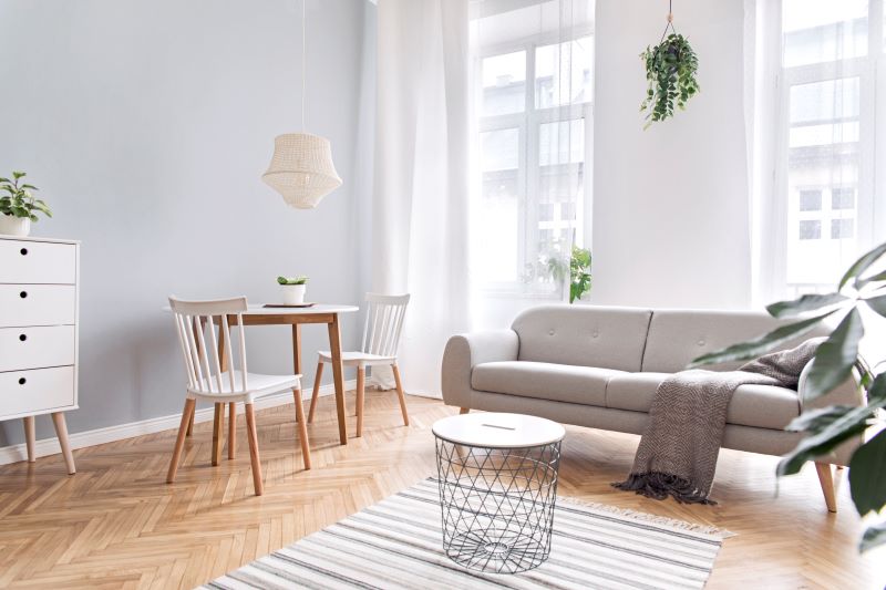 A minimalist living room with light blue walls, a gray sofa, a small dining table with two chairs, and a woven light fixture. The space is bright and airy with natural light filtering through large windows. Plants are placed around the room, adding greenery to the space. This design trick makes the small space feel bigger and brighter.