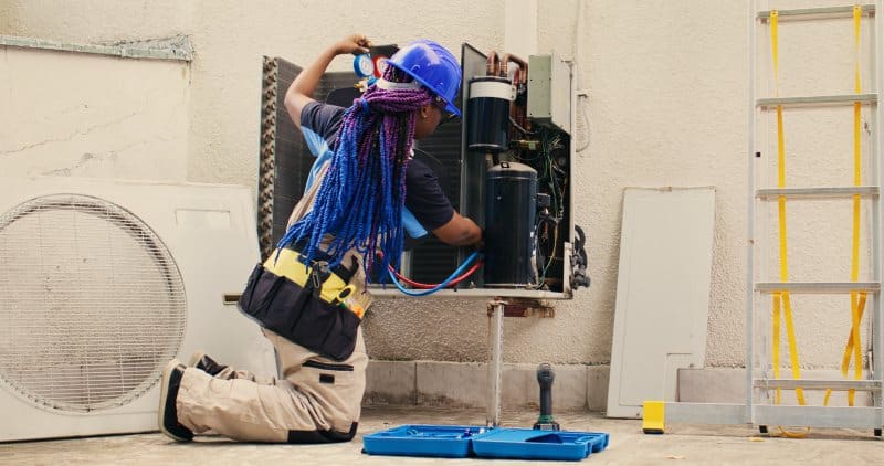 Technician performing maintenance on an air conditioning unit outdoors, wearing a blue helmet and tools belt, working with cooling system components.