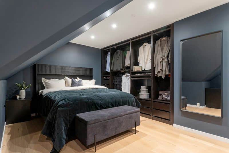 Modern bedroom with dark blue walls, open wardrobe, velvet bedding, and bench seating, showcasing stylish home decor in a cozy attic space.