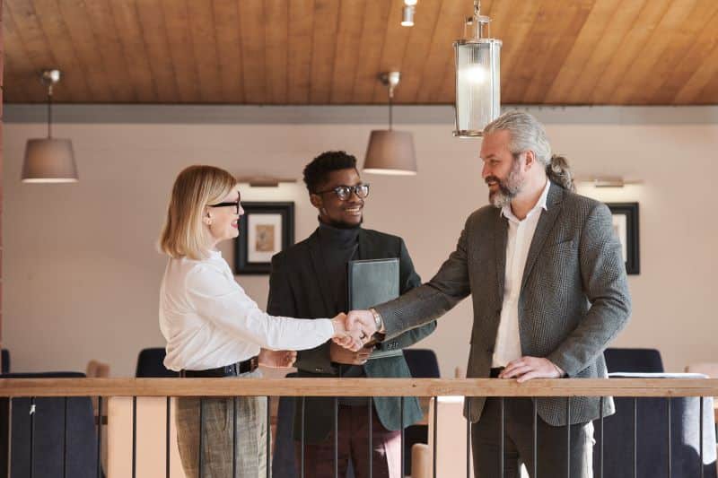 A woman and a man shaking hands while a third person observes. The scene takes place in a modern office setting, likely signifying a business deal or collaboration, a common practice among successful property investors.