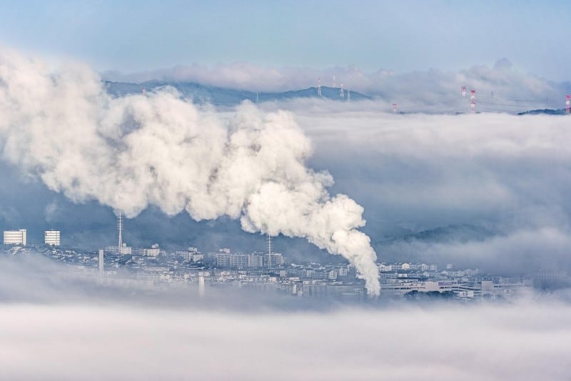 Thick industrial smoke rising from a city factory, blending into surrounding clouds, symbolizing carbon emissions and environmental impact.