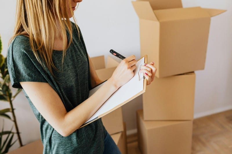Woman checking a moving checklist on a clipboard with stacked cardboard boxes in the background during a DIY relocation.