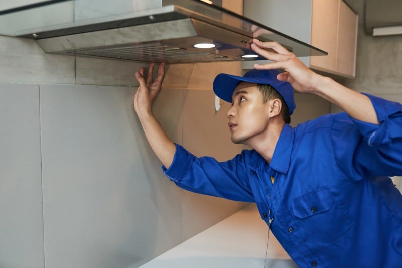 Technician in blue uniform inspecting a kitchen vent hood, checking for signs of buildup that may require professional duct cleaning.
