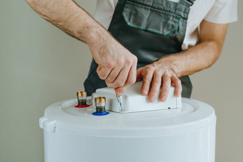 Technician installing or repairing a hot water system, using a screwdriver to secure the top panel near water inlet connections.