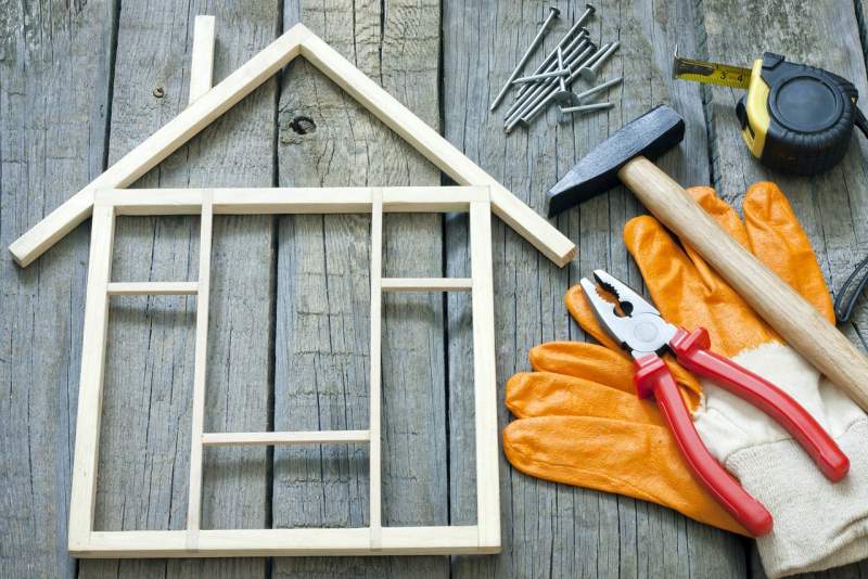 Wooden house frame outline with construction tools, nails, gloves, hammer, pliers, and tape measure on rustic wooden background for home renovation concept.