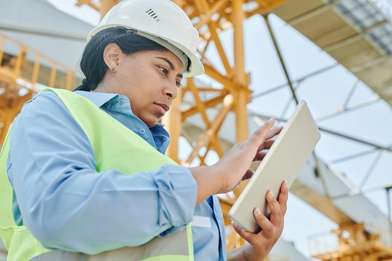 Female construction worker in safety vest and helmet inspecting lifting equipment site using a digital tablet during LOLER check.