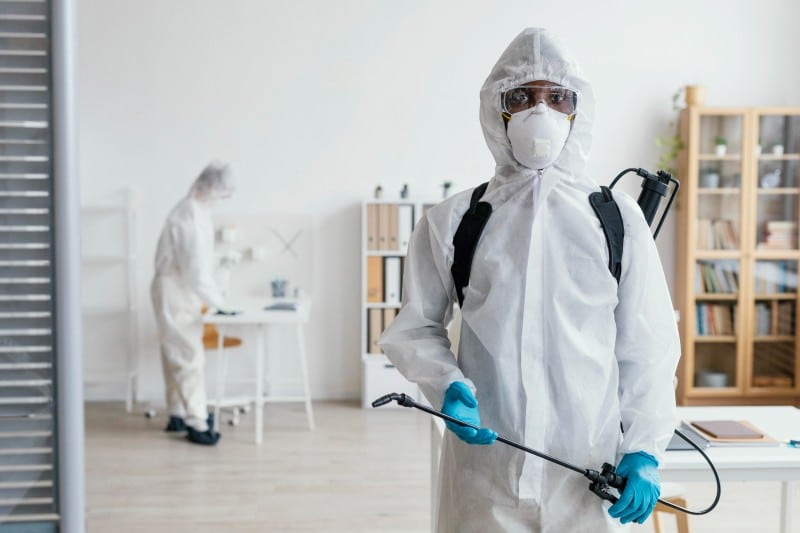 Person in protective suit using eco-friendly pest control sprayer indoors, ensuring family-safe treatment and clean living space.