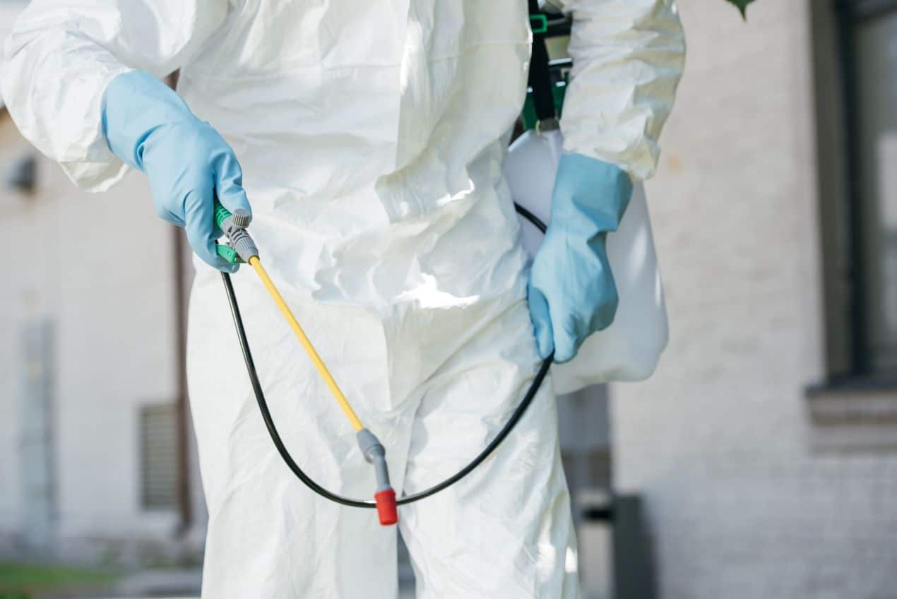 Professional technician in protective suit and blue gloves using a sprayer for pest control treatment outside a home.
