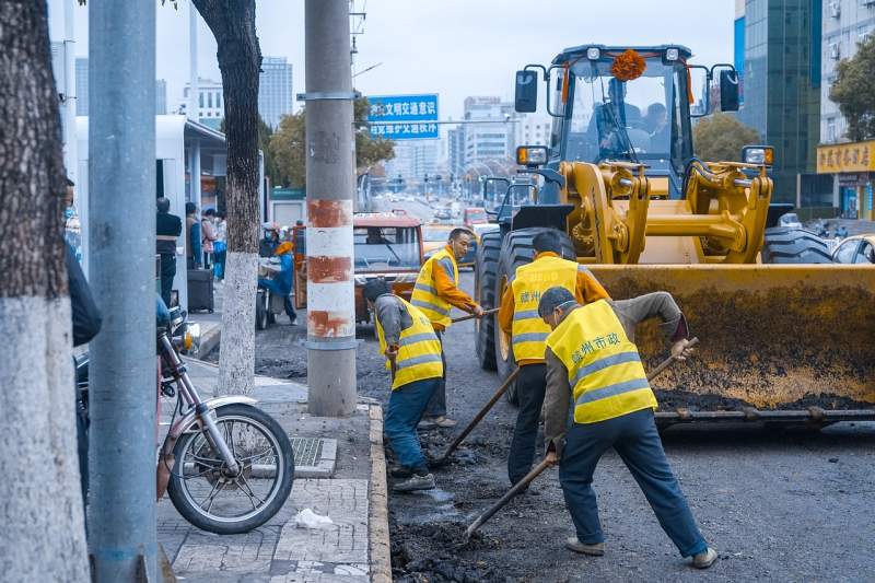 Workers in yellow safety vests use shovels to repair asphalt beside a loader, improving road durability through proper installation.