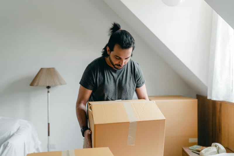 Man lifting a large cardboard box in a bedroom, organizing belongings to create more storage space.