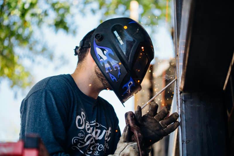 A man wearing a welding helmet and gloves performs welding on a metal structure, with sparks flying—demonstrating basic welding for home repairs.