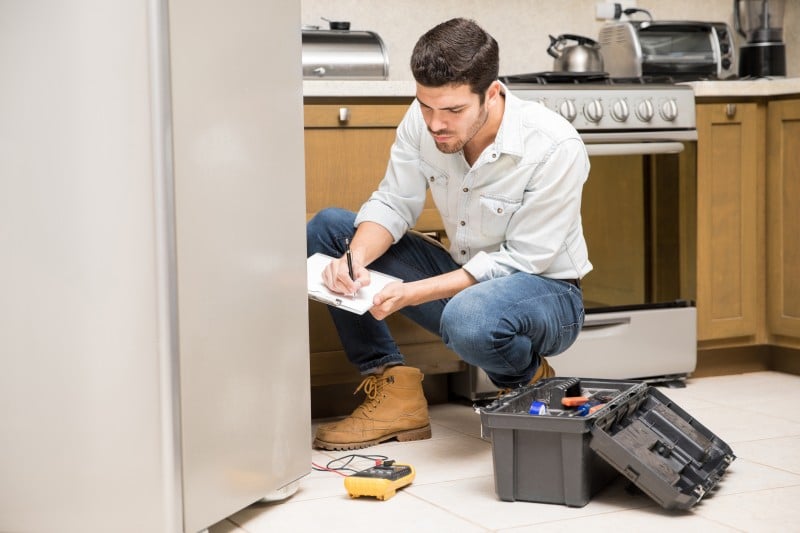 A technician kneeling in a kitchen, inspecting a refrigerator and writing notes on a clipboard with a toolbox nearby. Early inspection helps prevent appliance failure.