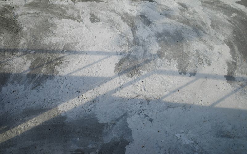 Close-up of a concrete floor being prepped, showing rough surface texture, dust, and partial grinding marks, with shadows from a railing cast across the surface.
