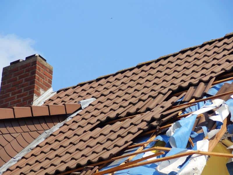 Close-up of a house roof with visible roof damage where tiles are broken and shifted, exposing wooden beams and blue tarp underneath. Example of roof damage needing replacement in Charlotte.