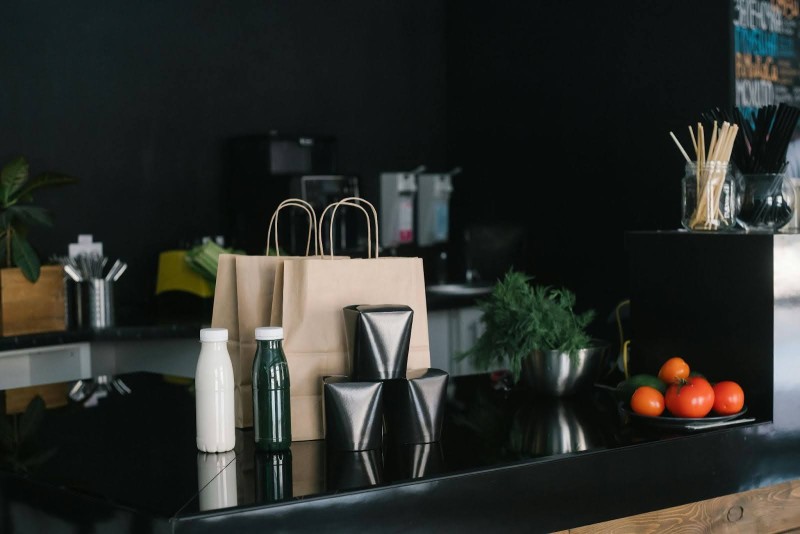Brown paper bags, reusable bottles, and fresh vegetables on a black countertop in a modern kitchen, highlighting eco-friendly and sustainable design trends.