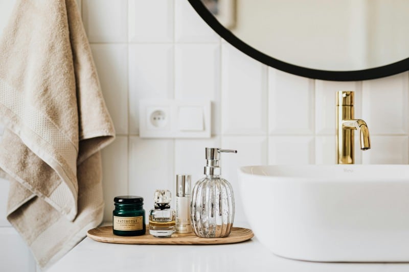 Modern bathroom with a white sink, gold faucet, toiletries on a wooden tray, and beige towels hanging by a round mirror. Family-friendly bathroom design.