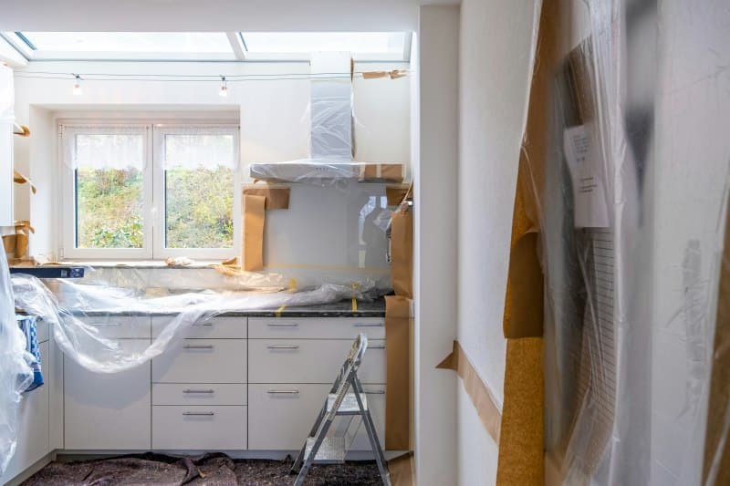Kitchen under renovation with cabinets and appliances covered in plastic, ladder in center, and light from skylight and window.