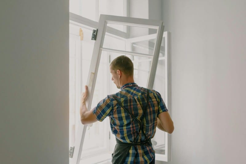 A worker in a plaid shirt and apron installing or adjusting a modern white-framed window inside a home, representing practical home upgrades that can add real value.