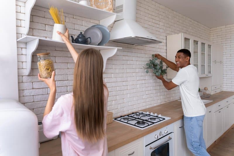 A woman stores pasta on a shelf while holding a jar, and a man decorates the wall with greenery in a modern white brick kitchen remodel.